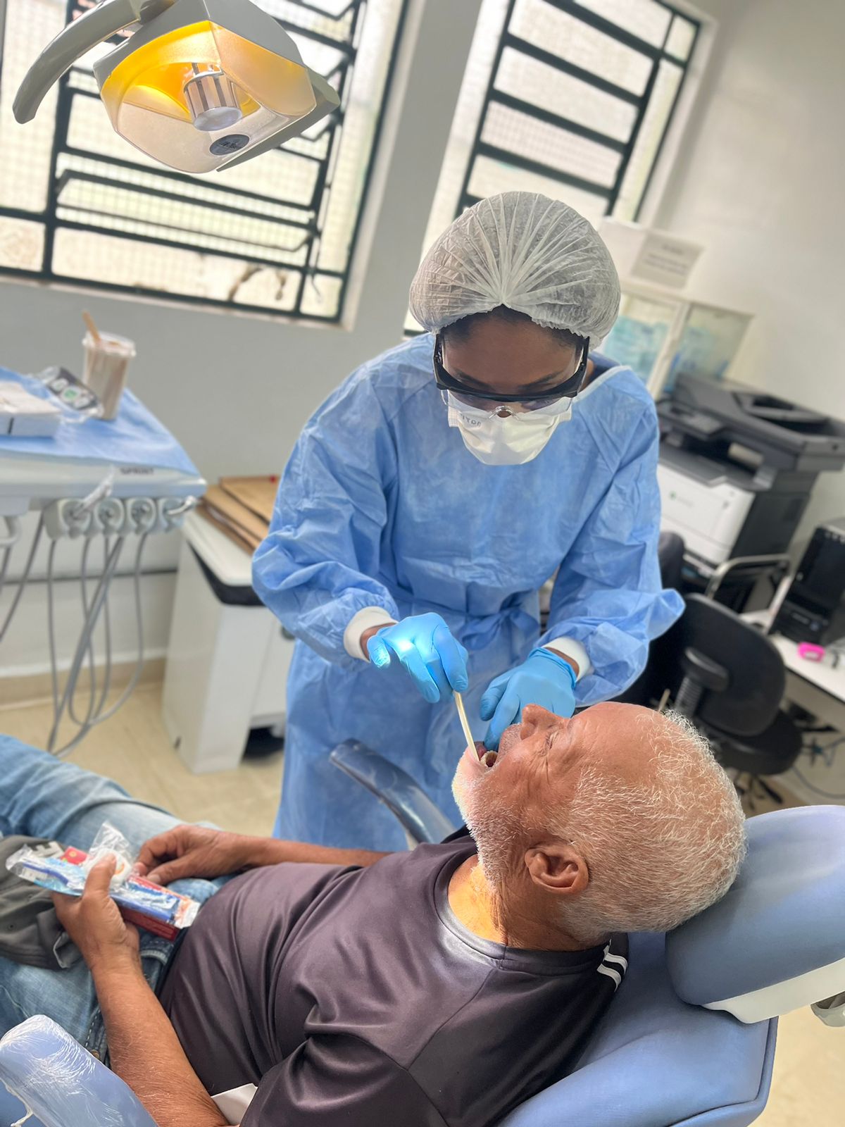 A foto mostra uma dentista durante um exame em paciente. Ela usa avental azul, touca, máscara e luvas, e usa uma espátula para examinar a boca do paciente, um homem idoso